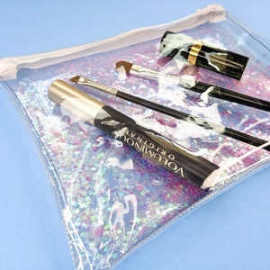 May include: A clear, iridescent cosmetic bag filled with makeup items. The bag contains a tube of mascara, two makeup brushes, and a lipstick. The mascara tube has the text "VOLUMINOUS ORIGINAL" printed on it.