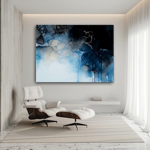 Op de afbeelding: Abstracte wandkunst met blauwe, zwarte en witte inktpatronen. Het kunstwerk hangt in een moderne woonkamer met een witte stoel en ottoman, en een neutraal tapijt.