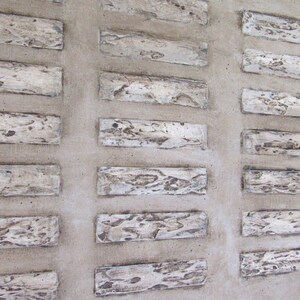 Op de afbeelding: Een close-up van een muur met een patroon van rechthoekige tegels. De tegels zijn gemaakt van een lichtgekleurd materiaal, mogelijk steen of beton, en hebben een ruw, getextureerd oppervlak. De tegels zijn gerangschikt in een rasterpatroon, waarbij elke tegel iets verschoven is ten opzichte van de volgende.