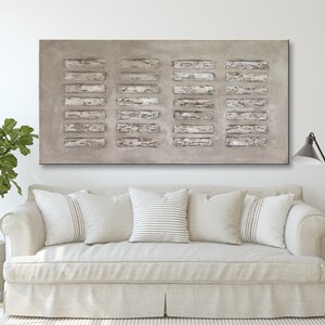 Op de afbeelding: Een groot abstract schilderij met een grijze achtergrond en een patroon van witte rechthoekige vormen. Het schilderij hangt boven een witte bank met kussens.
