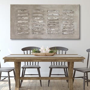Op de afbeelding: Een rustieke houten eettafel met vier grijze stoelen. De tafel heeft een gestreepte loper en een houten kom met groen en kaarsen in het midden. Een groot abstract schilderij in grijs en wit hangt aan de muur achter de tafel.
