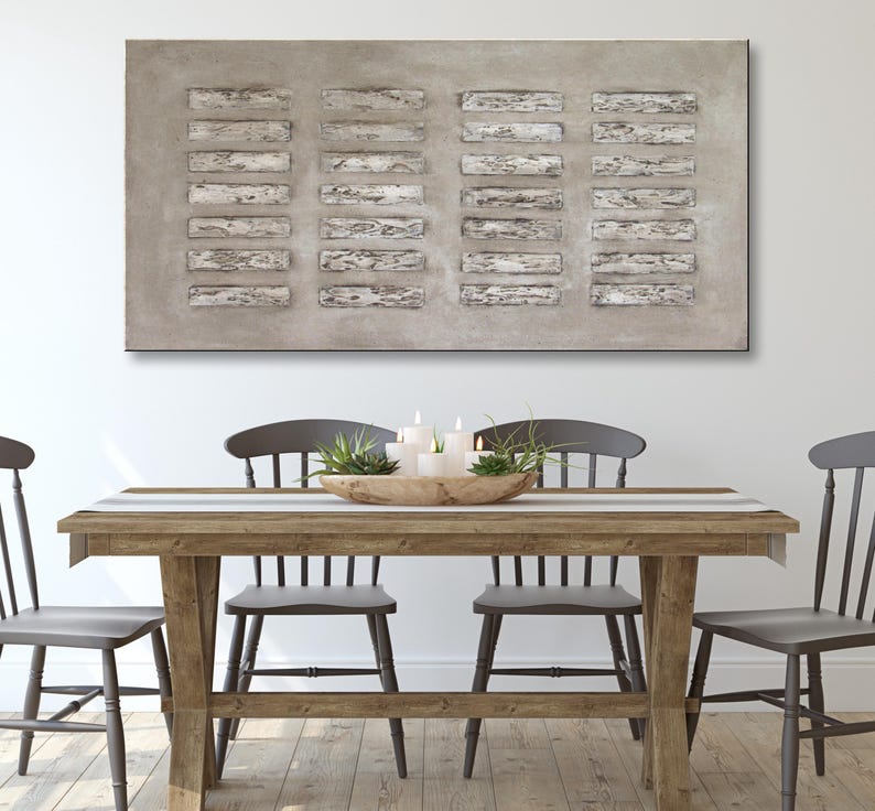 Op de afbeelding: Een rustieke houten eettafel met vier grijze stoelen. De tafel heeft een gestreepte loper en een houten kom met groen en kaarsen in het midden. Een groot abstract schilderij in grijs en wit hangt aan de muur achter de tafel.