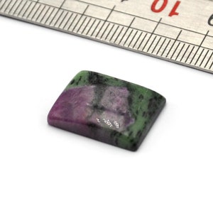 Può includere: Una piccola gemma quadrata con un mix di colori verdi, rosa e neri. Un righello di metallo è posto diagonalmente sopra la gemma. La gemma misura circa 2 cm di lunghezza.