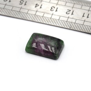 Può includere: Un cabochon di pietra preziosa verde e rosa di forma rettangolare, che misura circa 1 cm di lunghezza. La pietra preziosa è su una superficie bianca accanto a un righello.