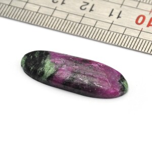 May include: A close-up of a green, pink, and black oval-shaped gemstone with a ruler for scale. The gemstone measures approximately 3.8 cm in length.