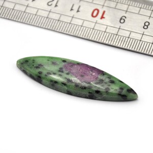 May include: A green and pink oval-shaped gemstone with black speckles. The gemstone is about 2 inches long and is sitting on a white surface next to a metal ruler.