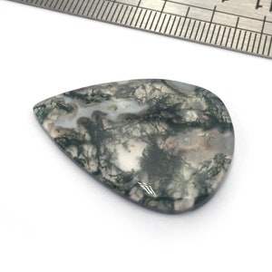 Peut inclure: Un cabochon de agate mousse verte en forme de larme sur fond blanc. La pierre mesure environ 5 cm de long et 2,5 cm de large.