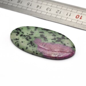May include: Oval-shaped gemstone with a green base, speckled with black and a prominent pinkish-red section. A metal ruler is positioned nearby, providing a size reference. The stone is set against a white background.