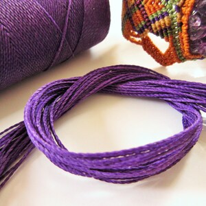 Könnte beinhalten: Lilafarbener Baumwollschnur, eine Spule mit der gleichen Schnur und ein lilafarbenes und orangefarbenes Makramee-Armband mit Amethyststeinen.