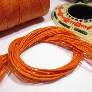 Könnte beinhalten: Nahaufnahme einer Spule mit orangefarbenem Garn und einem Stapel orangefarbenen Garns. Das Garn ist dünn und hat einen leichten Glanz.