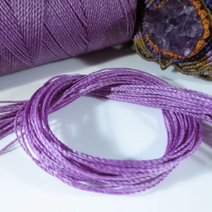 Könnte beinhalten: Nahaufnahme eines geflochtenen violetten Kordels, das die Textur und die Details des Kordels zeigt. Der Kordel ist zu einem Kreis gewickelt und es hängen lose Stränge von der Spule.