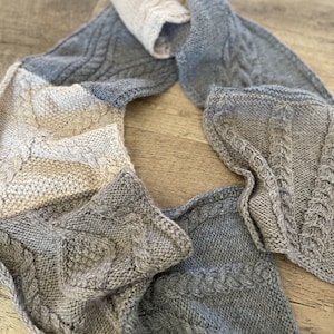 May include: A knitted scarf in shades of gray and beige with a cable knit pattern.