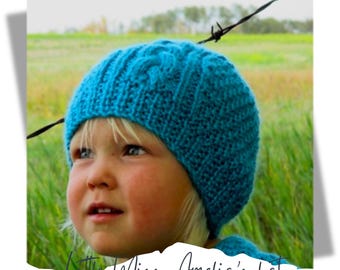 May include: A close-up of a child wearing a blue knitted hat with a cable stitch pattern. The hat is pulled down over the child's ears and forehead. The child is looking up and smiling. The background is a field of green grass.