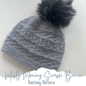 May include: A gray knitted beanie with a textured pattern and a fluffy, dark gray pom-pom on top. The text "Misty Morning Simple Beanie Knitting Pattern" is at the bottom of the image.