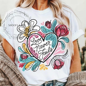 May include: White t-shirt with a colorful floral design and a heart with the text "Love Never Fails" in black lettering.
