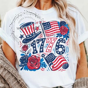 May include: White t-shirt with a red, white, and blue graphic design featuring the number 1776, stars, flags, and roses.