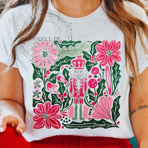 May include: White t-shirt featuring a colorful design with a nutcracker, flowers, and foliage in pink, green, and white. The design has a festive, holiday theme. The shirt is made of a soft material.