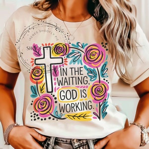 May include: A white t-shirt with a colorful floral design and a white cross. The text "In the waiting, God is working." is printed on the shirt.