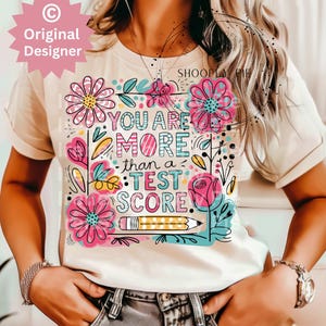 May include: A white t-shirt with a colorful graphic design featuring flowers, a pencil, and the text "You are more than a test score."