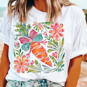 May include: White t-shirt with a colorful carrot and floral design. The carrot is tied with a pink and green bow. The flowers are pink, yellow, and green.