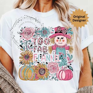 Op de afbeelding: Wit T-shirt met een kleurrijk grafisch ontwerp met een vogelverschrikker, pompoenen, zonnebloemen en de tekst "Too Fab for Flannel". Het ontwerp bevat roze, blauwe, gele en oranje elementen, perfect voor de herfst.