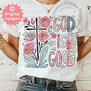 以下が含まれることがあります： 白いTシャツに花柄のデザインと「God is good」というピンクと黒の文字が書かれています