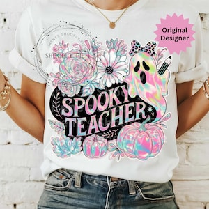 May include: White t-shirt with a colorful "Spooky Teacher" graphic. The design features a ghost, pumpkins, flowers, and the text "Spooky Teacher". The ghost has a bow and pencil. The shirt has rolled-up sleeves.