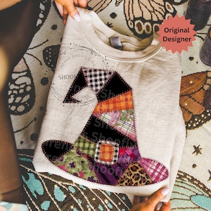 Könnte beinhalten: Ein hellgraues Sweatshirt mit einem Patchwork-Hexenhut-Design. Der Hut besteht aus verschiedenen gemusterten Stoffen, darunter Karos, Blumen und Leopardenmuster. Der Text "SHOOFY PIE DESIGNS" ist auf das Shirt gedruckt. Ein roter Stern mit den Worten "Original Designer" ist ebenfalls sichtbar.