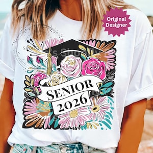 以下が含まれることがあります： 卒業デザインのフローラルデザインが施された白いTシャツ。デザインには卒業帽子、バラ、デイジーが含まれています。「SENIOR 2026」と書かれたバナー。デザインにはピンク、白、ターコイズの色が含まれています。「Original Designer」の文字も表示されています。