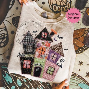 Könnte beinhalten: Beiges Sweatshirt mit einem bunten Patchwork-Haus-Design mit Fledermäusen und einem Halbmond. Die Häuser haben verschiedene Muster, darunter Blumen-, Karo- und Leopardenmuster. Der Text "Irenes Shoofly Pie" ist auf dem Sweatshirt aufgedruckt.