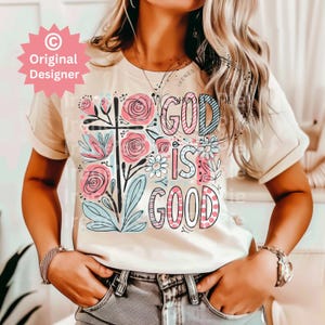以下が含まれることがあります： 花柄のデザインと「God is good」という文字がピンク、ブルー、ブラックで書かれた白いTシャツです。