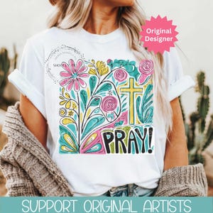 May include: White t-shirt featuring a colorful floral design with a yellow cross and the word "PRAY!" in bold letters. The design includes pink, blue, and yellow flowers and leaves. The shirt has rolled-up sleeves.