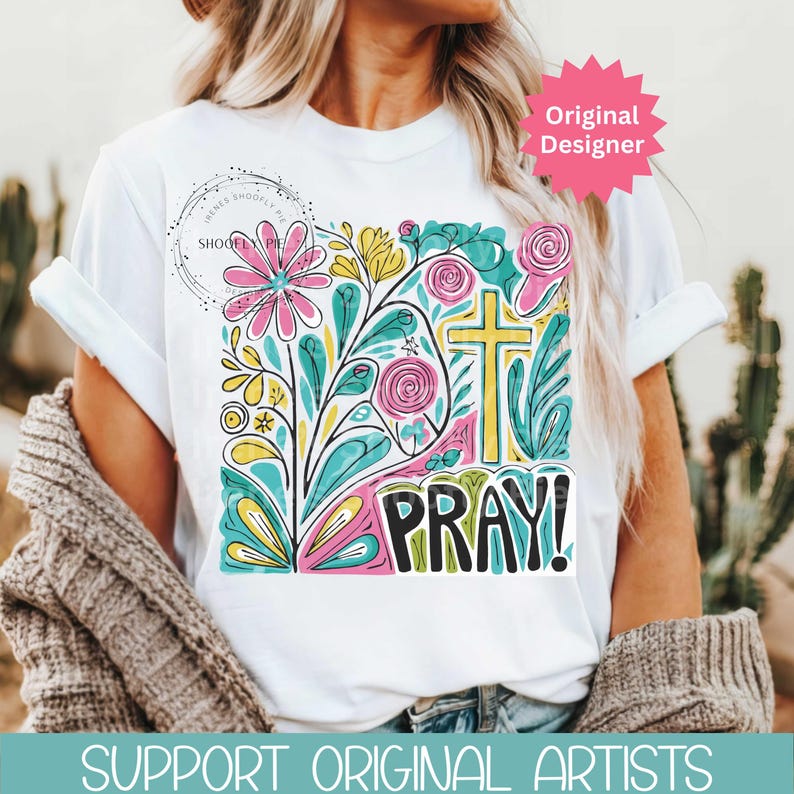 May include: White t-shirt featuring a colorful floral design with a yellow cross and the word "PRAY!" in bold letters. The design includes pink, blue, and yellow flowers and leaves. The shirt has rolled-up sleeves.