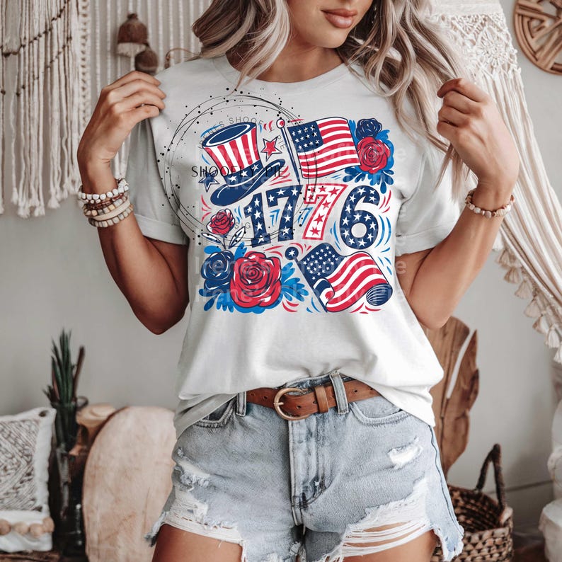 May include: White t-shirt with a red, white, and blue graphic design featuring a top hat, stars, and the number 1776. The graphic also includes red roses and American flags.