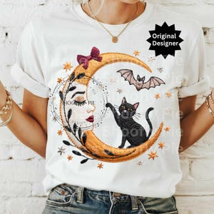Op de afbeelding: Wit T-shirt met een geborduurd ontwerp met een maan, een zwarte kat en een vleermuis. De maan heeft een gezicht en een strik. Het ontwerp bevat sterren en de tekst "SHOOGY PIE DESIGN".