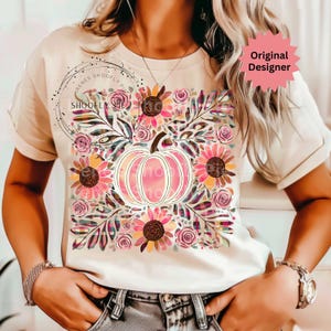 May include: Cream-colored t-shirt featuring a floral and pumpkin design. The graphic includes pink sunflowers, roses, and a pink pumpkin. The text "SHOOFly" is visible in a circular design. The words "Original Designer" are also visible.