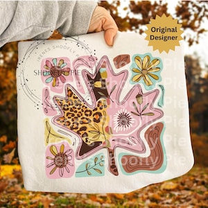 May include: A white square pillow featuring a large pink leaf design with floral and leopard print accents. The design includes flowers, an acorn, and the text "Irenes Shoofly Pie". The pillow is held by a person.