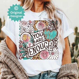 May include: White t-shirt with a floral design and the text "You Are Enough" in black lettering. The design includes a turquoise heart with a distressed look.