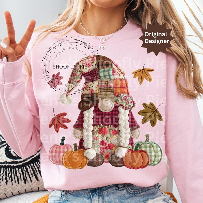 Peut inclure: Sweat-shirt rose clair avec un motif de gnome d'automne. Le gnome porte un chapeau patchwork, des d&eacute;tails floraux et une barbe blanche tress&eacute;e. Des citrouilles et des feuilles sont &eacute;galement repr&eacute;sent&eacute;es. Le texte "SHOOFY PIE" est visible.
