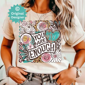 May include: A white t-shirt with a floral design and the text "You Are Enough" in black lettering. The design also includes a heart with a turquoise and brown pattern.