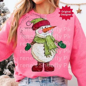 Op de afbeelding: Een roze sweatshirt met een grillig sneeuwpopontwerp. De sneeuwpop heeft een wortelneus, een groene sjaal, een rode hoed en groene wanten. De woorden "SHOONPIE" en "Original Designer" zijn ook zichtbaar.