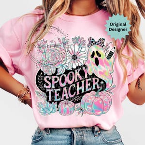 May include: Pink t-shirt with a colorful "Spooky Teacher" graphic. The design features a ghost, pumpkins, flowers, and the text "Spooky Teacher" in a black oval. The shirt has short sleeves.