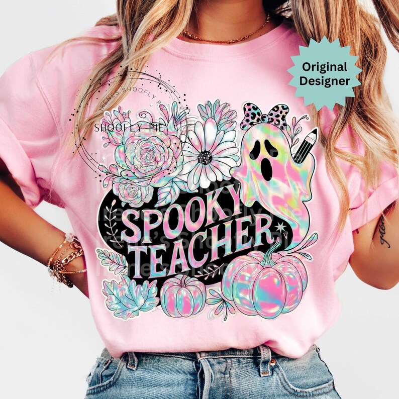 May include: Pink t-shirt with a colorful "Spooky Teacher" graphic. The design features a ghost, pumpkins, flowers, and the text "Spooky Teacher" in a black oval. The shirt has short sleeves.