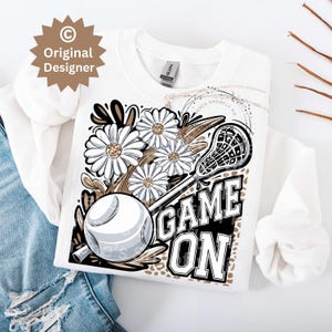 May include: White t-shirt with a graphic design featuring a lacrosse stick, a softball, daisies, and the text "GAME ON" in black and white.