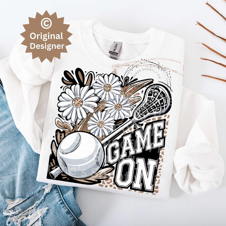 May include: White t-shirt with a graphic design featuring a lacrosse stick, a softball, daisies, and the text "GAME ON" in black and white.