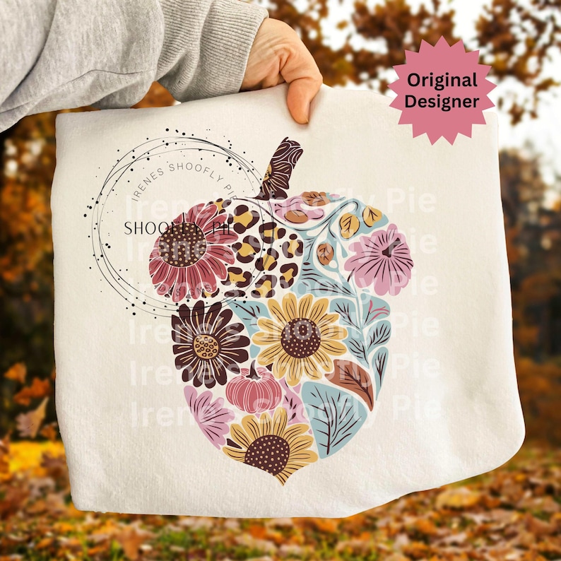May include: A cream-colored decorative pillow featuring a floral design in the shape of an acorn. The design incorporates sunflowers, leopard print, and other flowers in shades of pink, yellow, and brown. The text "Irenes Shoofly Pie" is printed on the pillow.
