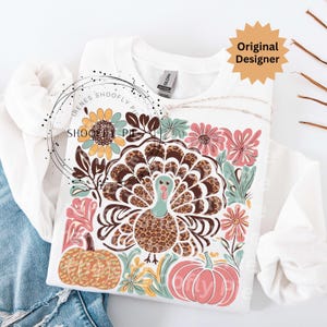 May include: White sweatshirt featuring a colorful Thanksgiving-themed design. The design includes a turkey with a leopard print pattern, pumpkins, and floral elements. The text "Original Designer" is visible on the top right.