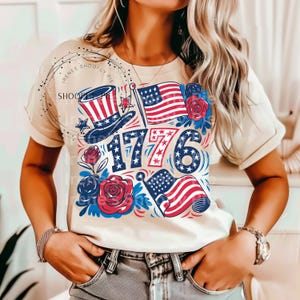 May include: A white t-shirt with a red, white, and blue graphic design featuring a liberty bell, stars, and the number 1776. The graphic also includes a red, white, and blue American flag.