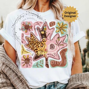 May include: White t-shirt featuring a large, colorful leaf design with floral and leopard print patterns. The design includes the text "Irenes Shoofly Pie" in a circular arrangement. The shirt also has a "Original Designer" tag.