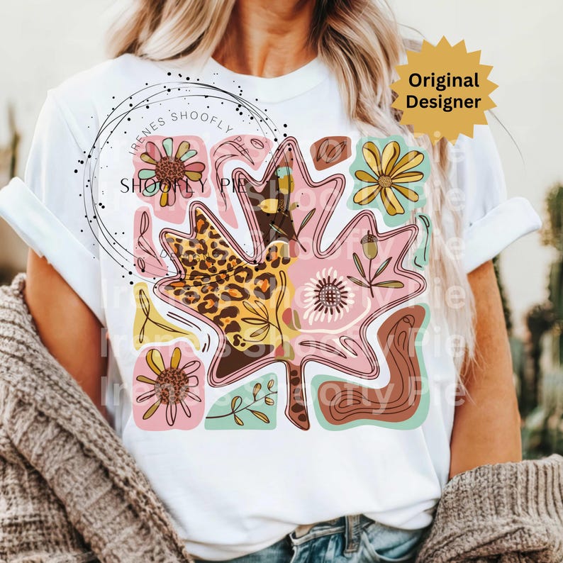 May include: White t-shirt featuring a large, colorful leaf design with floral and leopard print patterns. The design includes the text "Irenes Shoofly Pie" in a circular arrangement. The shirt also has a "Original Designer" tag.
